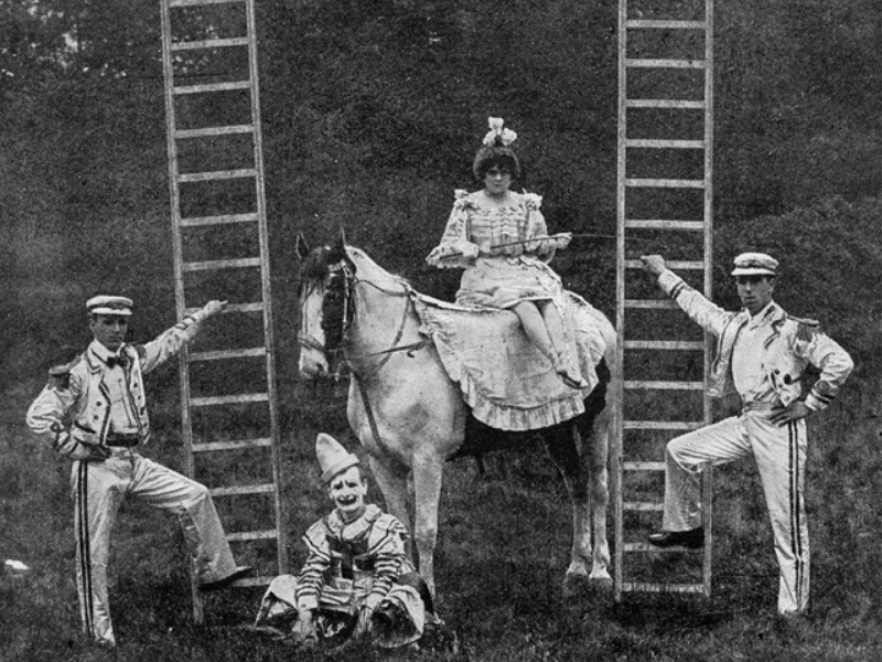 Circus scene with a woman in a frilly dress sitting on a horse. Two men in striped uniforms stand by ladders, and a clown sits cheerfully on the grass.