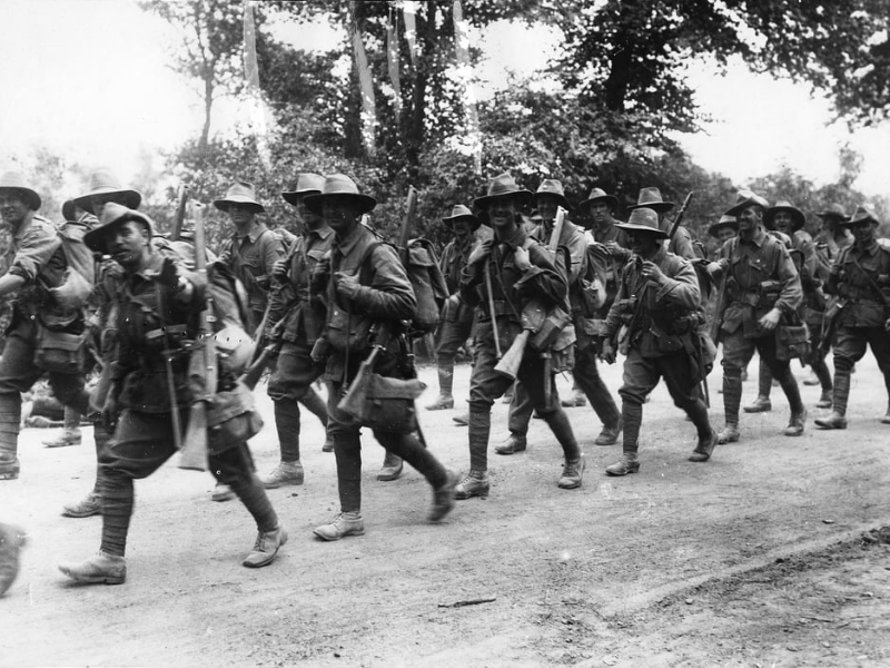 Group of World War I soldiers march along a tree-lined road, wearing broad-brimmed hats and carrying rifles. The scene conveys camaraderie and determination.