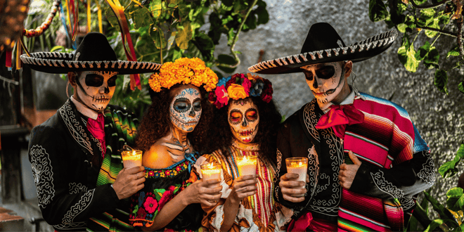 Four individuals dressed in traditional Day of the Dead attire, holding candles and gazing somberly at the flames, during a Mexican cultural celebration