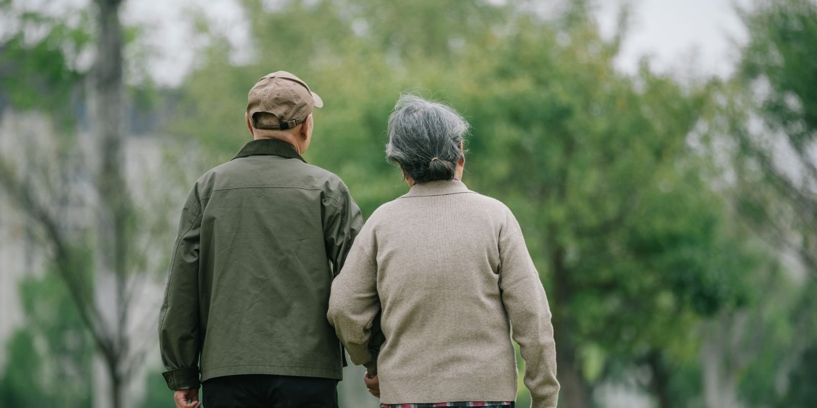 a male and female elderly couple walking through a green lush park on a soft cloudy day