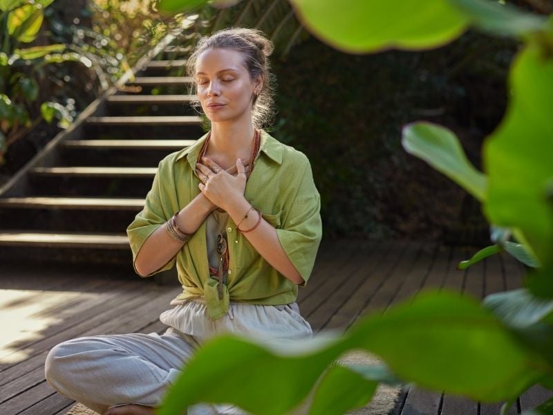 A woman in a green shirt sits cross-legged on a wooden deck, hands on chest, eyes closed, surrounded by lush plants, conveying calm and serenity.