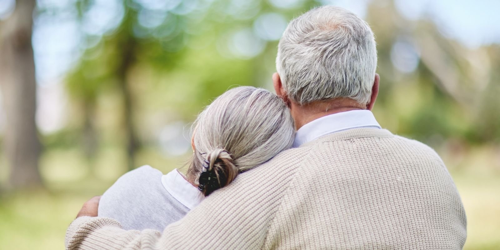 An elderly couple sits closely, embraced, with their backs to the camera in a lush, green park. The mood is peaceful and comforting under a soft, natural light.
