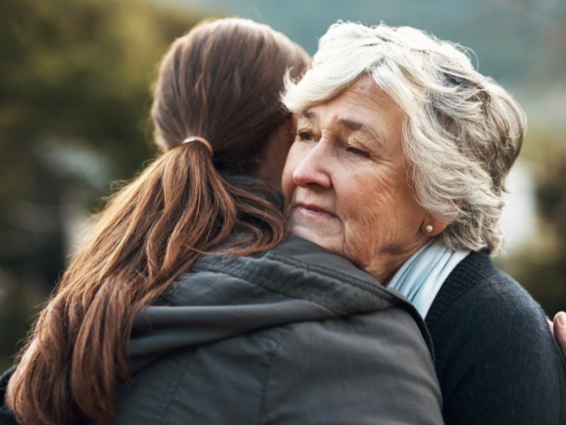 An elderly woman with white hair hugs a younger person outdoors. Her expression is serene yet contemplative, conveying warmth and affection amidst a blurred nature background.