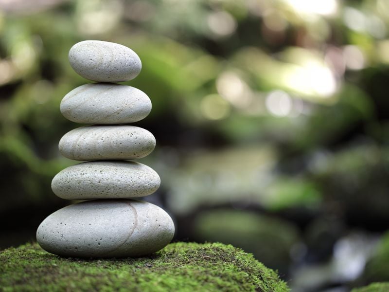 A stack of five smooth, gray stones balances on a mossy surface, set against a soft-focus green background creating a serene, tranquil atmosphere.