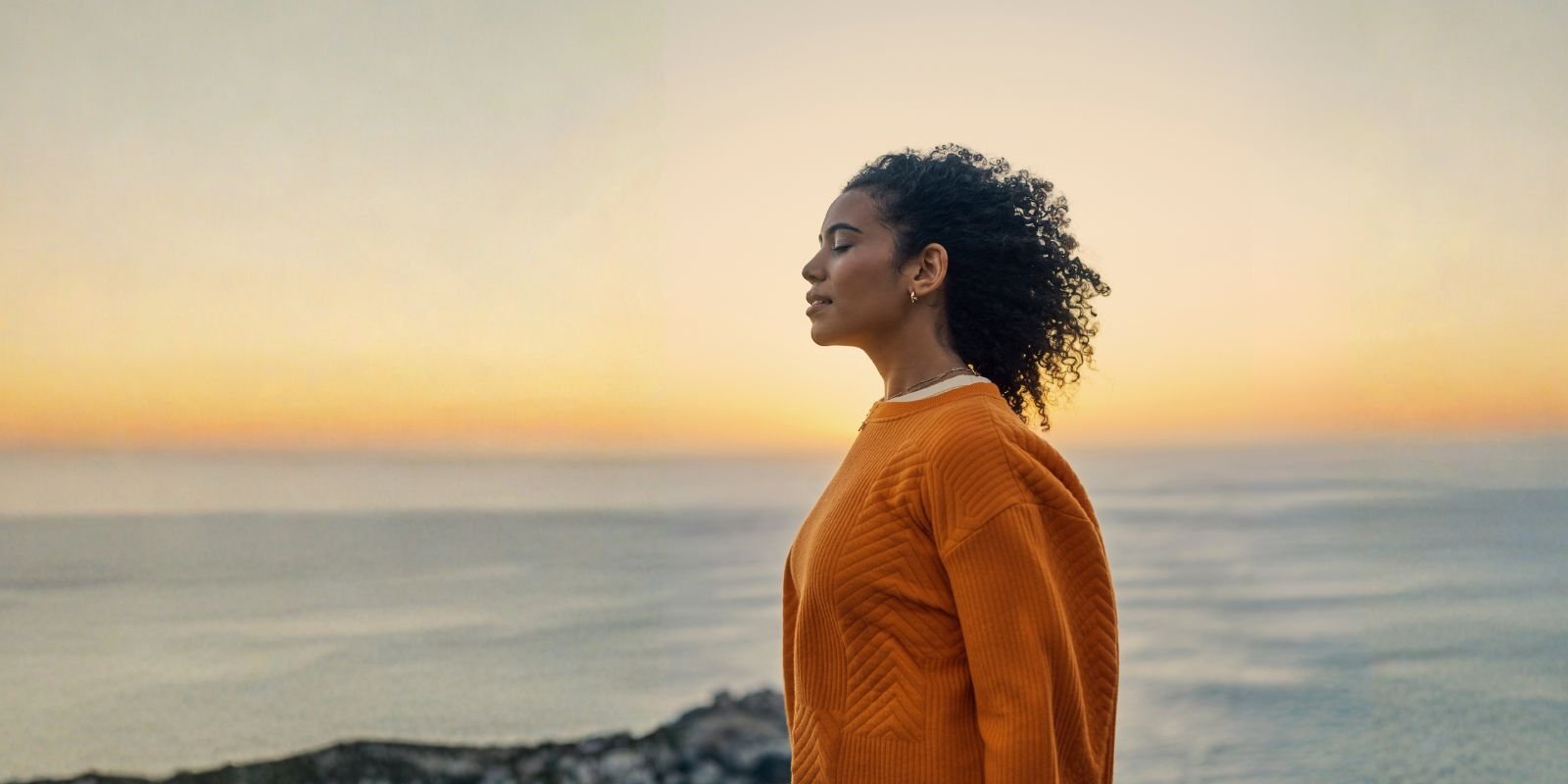 Girl closing eye and facing the ocean while the sun is setting.