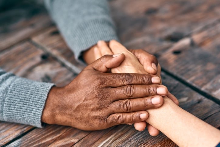 Two hands gently hold another hand on a wooden table, conveying comfort and support. The scene exudes warmth and empathy, emphasising human connection.