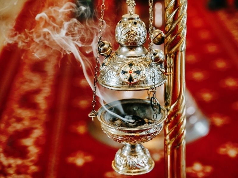 A silver incense censer hanging by chains, releasing wisps of smoke. The ornate design features intricate engravings and a decorative cross emblem. The background shows a rich red patterned carpet, creating a warm and reverent atmosphere.