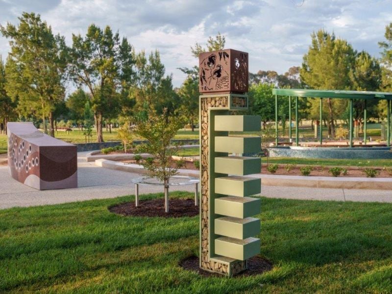 A geometric ash memorial stands on a green lawn in memorial park, surrounded by trees. A wavy bench and gazebo are nearby, under a partly cloudy sky.