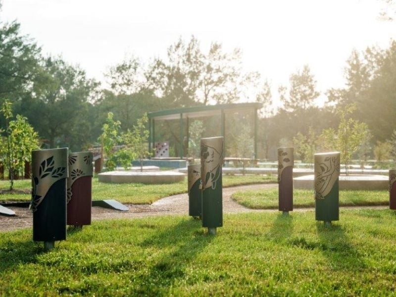 Sunlit memorial park scene with metal ash memorial garden totem posts featuring leaf designs, set on a grassy lawn. Pathways lead to a gazebo surrounded by trees. Calm atmosphere.