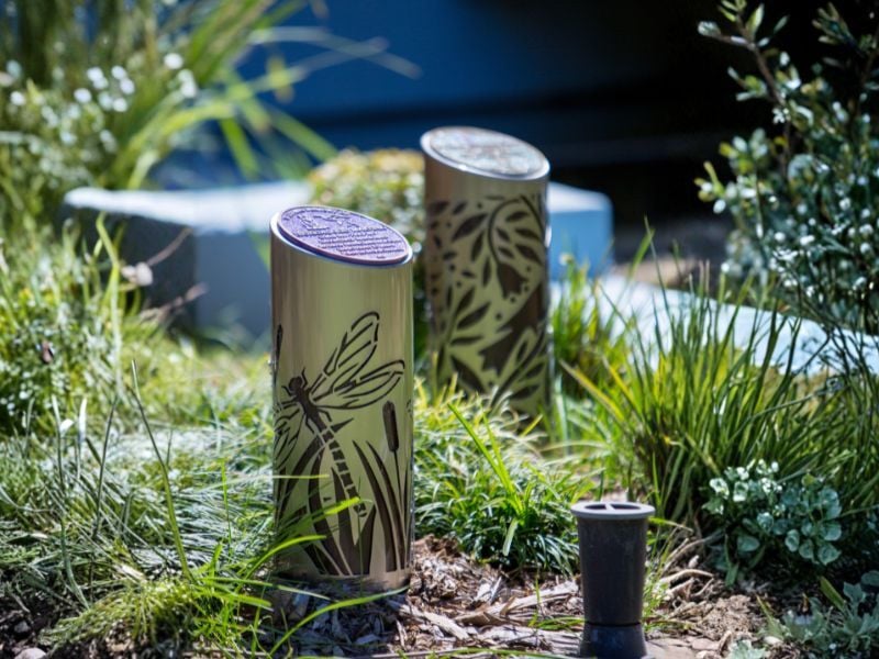 Two cylindrical ash memorial totem with dragonfly cutouts stand amidst lush greenery, creating a serene garden scene under bright sunlight.