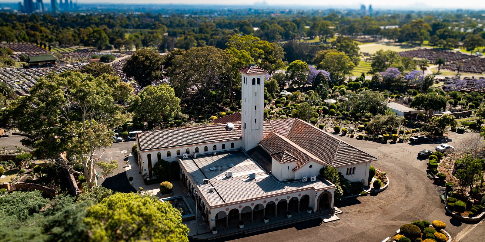 Aerial view of a grand white building with a crematorium, surrounded by lush greenery and gardens of Rookwood Crematorium. The scene conveys serenity with distant cityscape.