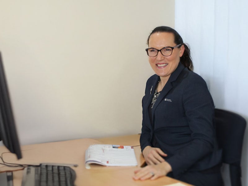 A woman in a Metropolitan Memorial Parks navy blazer sits at a desk with a computer and open planner. She is smiling, conveying a professional and friendly demeanour.