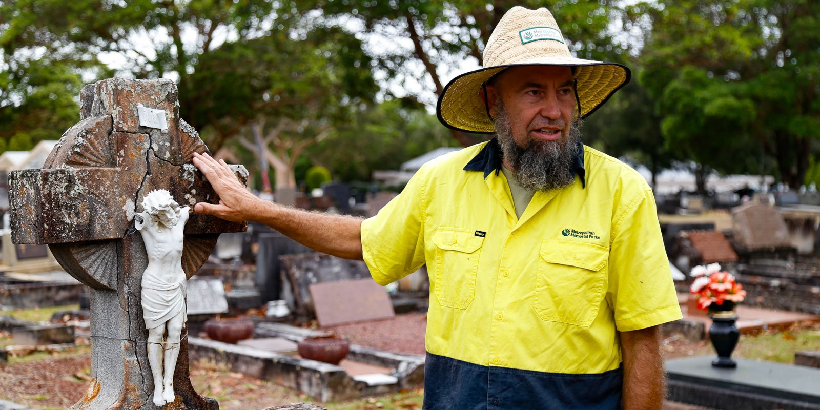 Metropolitan Memorial Parks restoration supervisor with straw hat stands at a cemetery, touching a weathered gravestone adorned with a cross and statue.
