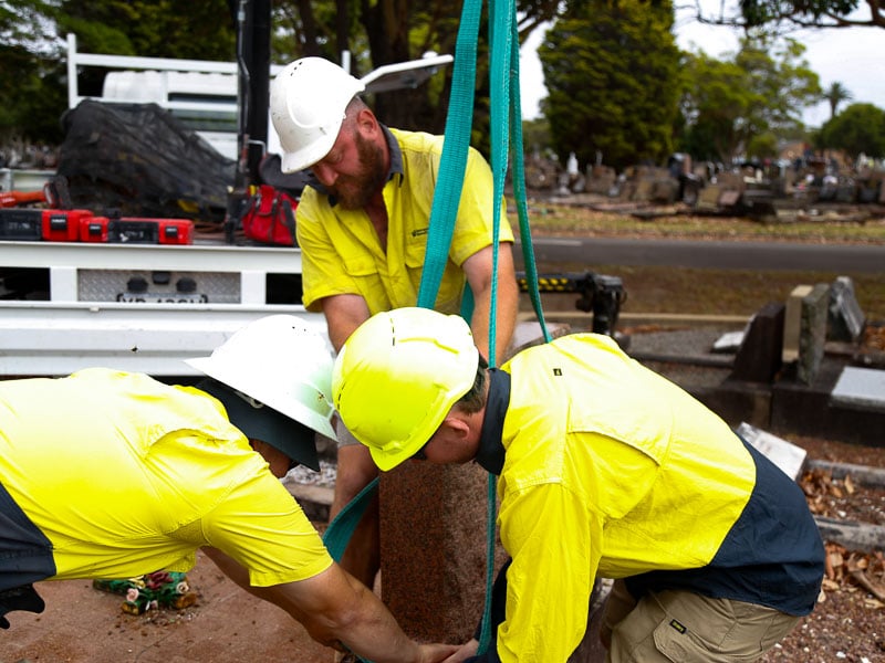 Metropolitan Memorial Parks monument restoration team in uniform and helmets are using straps to lift a heavy object in a cemetery. Tools are also visible on a nearby truck.