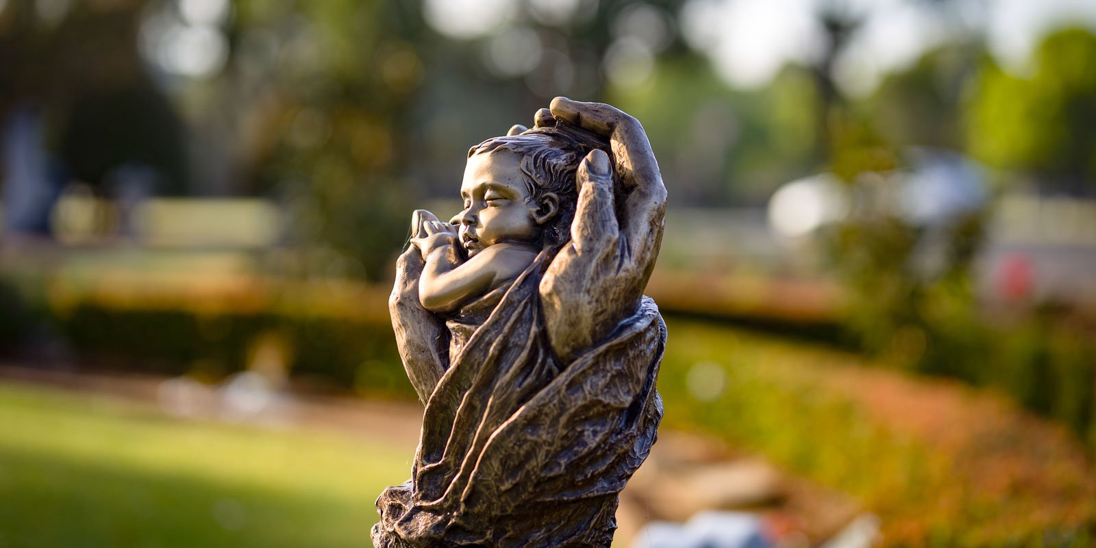 Bronze sculpture of hands cradling a baby, set in a peaceful garden with soft, blurred greenery at Woronora Memorial Park in the background.