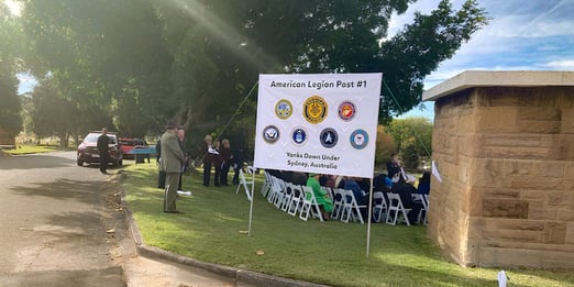 People gathering outdoors under trees for a ceremony, seated in white chairs. A banner for "American Legion Post #1" is displayed nearby.