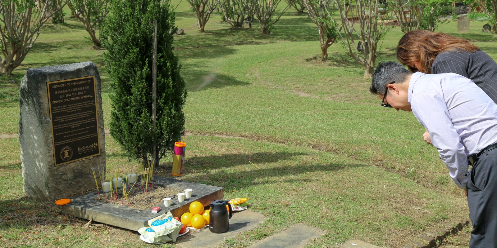 A man and woman bow in respect at a memorial stone for Chinese gold miners. Offerings of incense, candles, fruit, and drinks are placed nearby. The setting is a serene cemetery with neatly trimmed grass and trees at Rookwood General Cemetery.