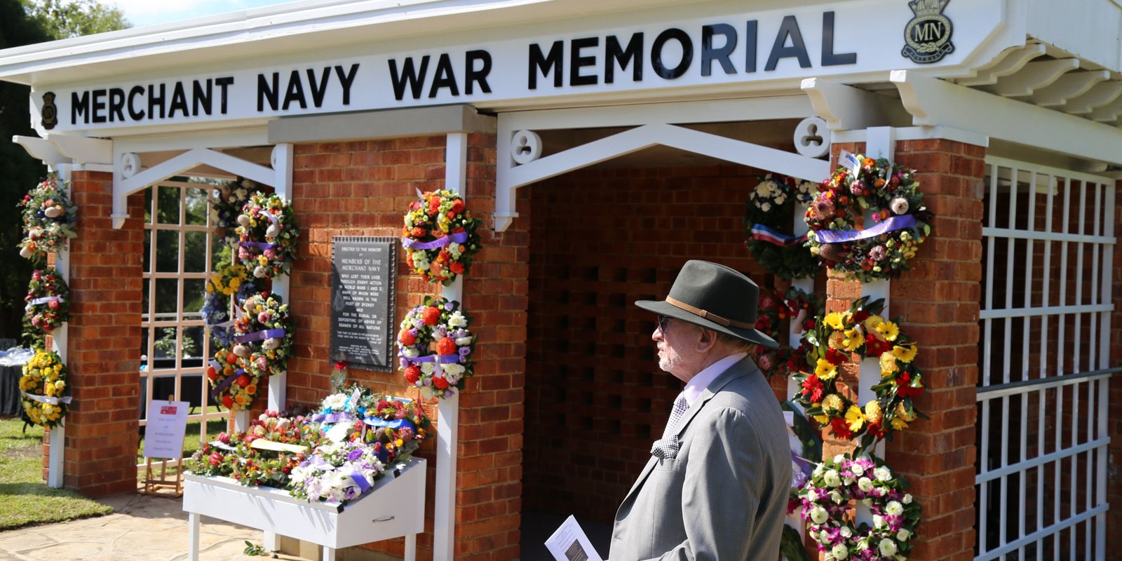 A man in a suit and hat stands solemnly before the brick Merchant Navy War Memorial adorned with colourful flower wreaths and a plaque.
