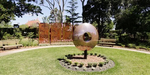 A round reflective metal sculpture with a circular hole sits in a vibrant garden. Two benches and decorative orange panels are surrounded by lush trees and greenery.