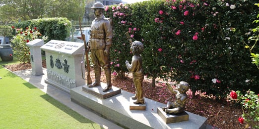 Bronze statues of an ANZAC soldier, a standing child, and a seated child on a stone platform surrounded by bushes and flowers. The standing child is handing a note to the soldier.