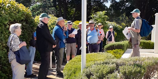 A group of people wearing casual outdoor clothing and hats, listen attentively to a tour guide with a backpack, next to a flagpole at Woronora Memorial Park