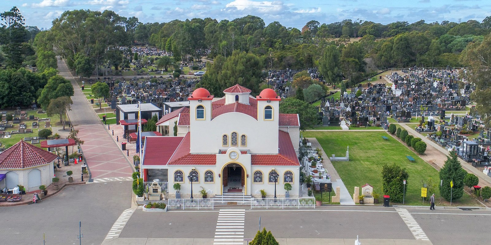Aerial view of a cemetery with a red-roofed Greek Orthodox church in the foreground. Rows of headstones are visible, surrounded by trees and pathways.