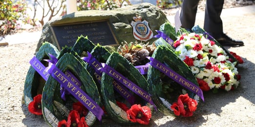 Ceremonial wreaths with "Lest We Forget" ribbons are placed in front of a memorial plaque on a stone. Red poppies and roses add solemnity and respect.