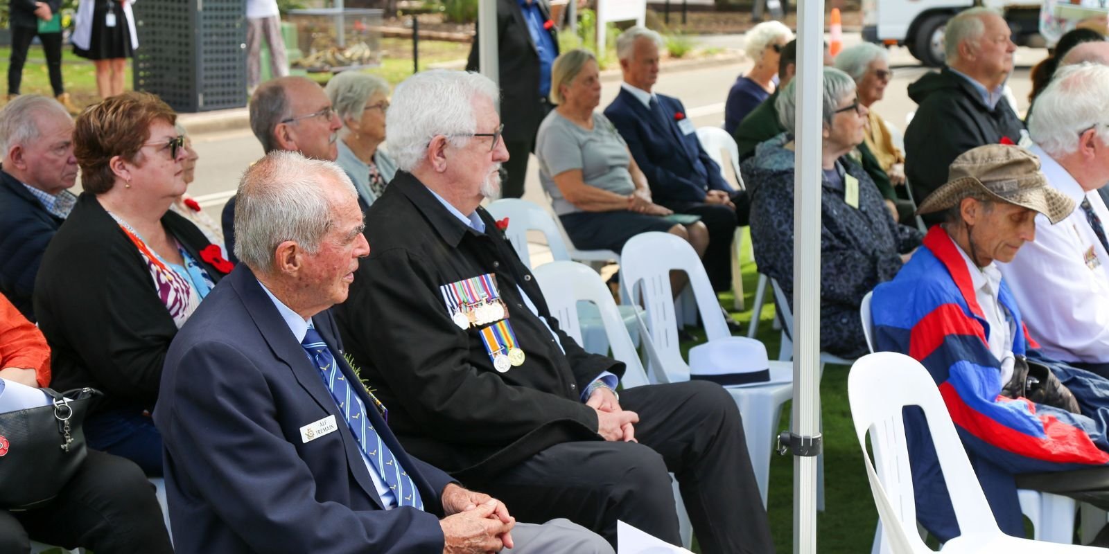 A diverse group of people, including veterans with medals, sit in white chairs at an outdoor commemorative event, some wearing red poppies.