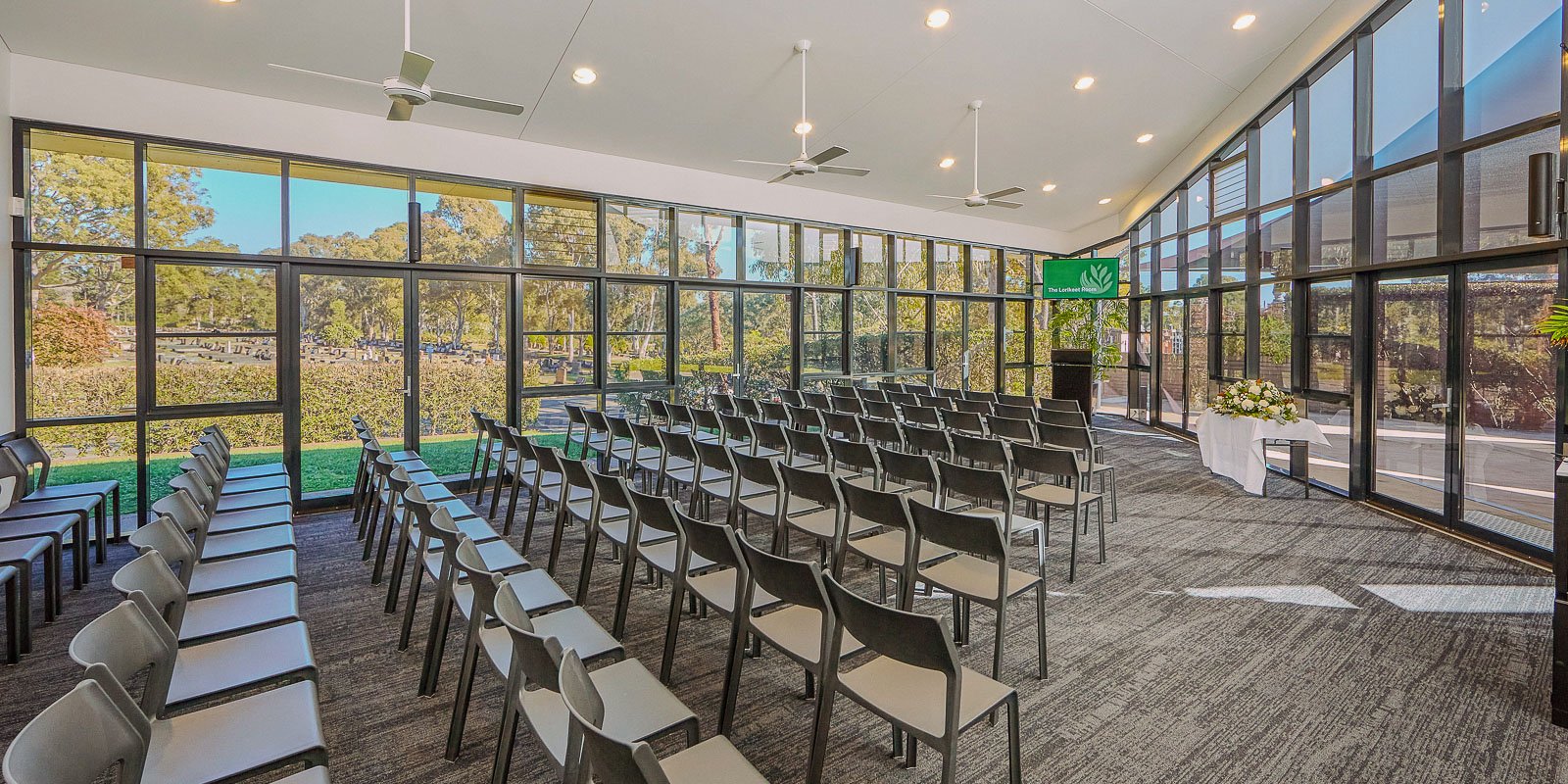 Bright chapel with large floor-to-ceiling windows, rows of chairs, and a view of trees and greenery outside.