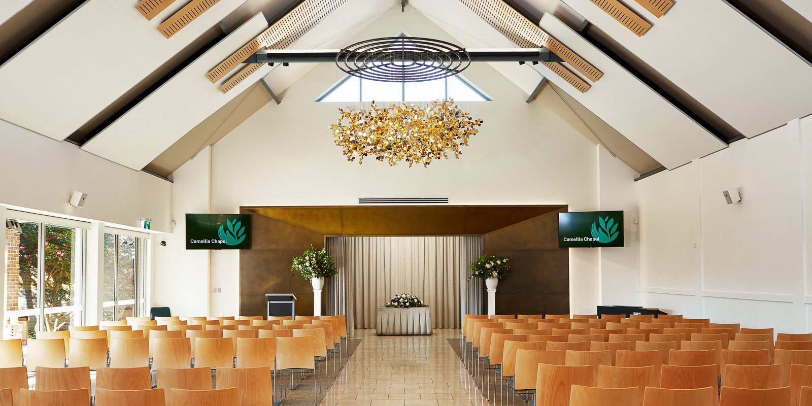 Modern chapel interior with high ceilings, wooden chairs, a floral chandelier, and a stage at the front.