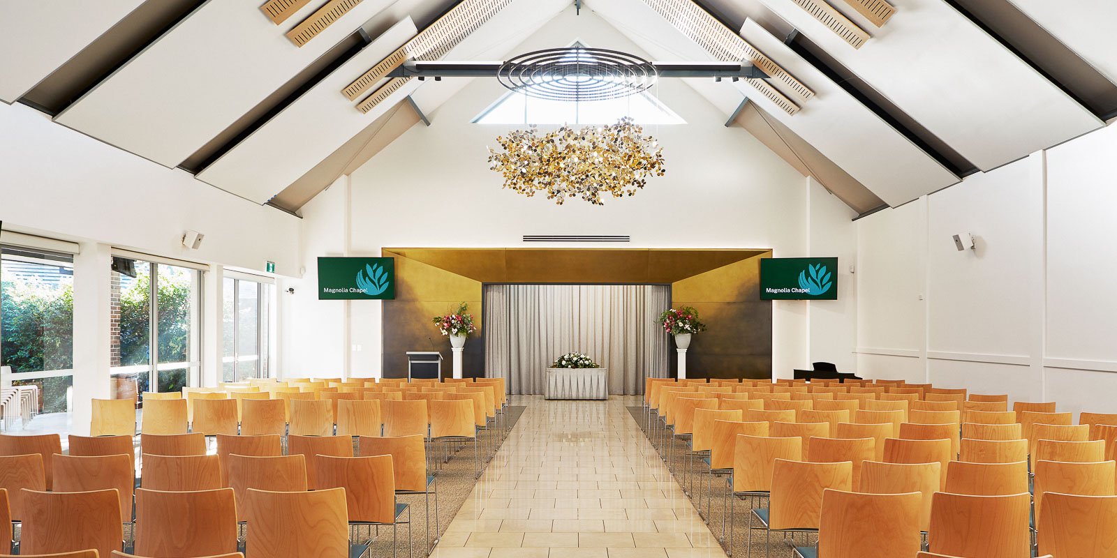 Modern chapel interior with high ceilings, wooden chairs, a floral chandelier, and a stage at the front.
