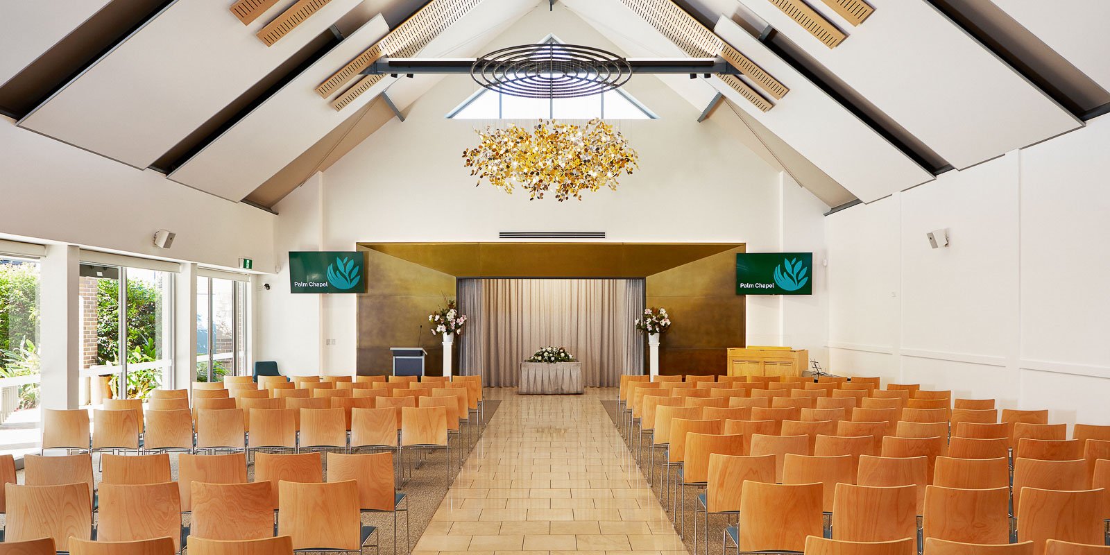 Modern chapel interior with high ceilings, wooden chairs, a floral chandelier, and a stage at the front