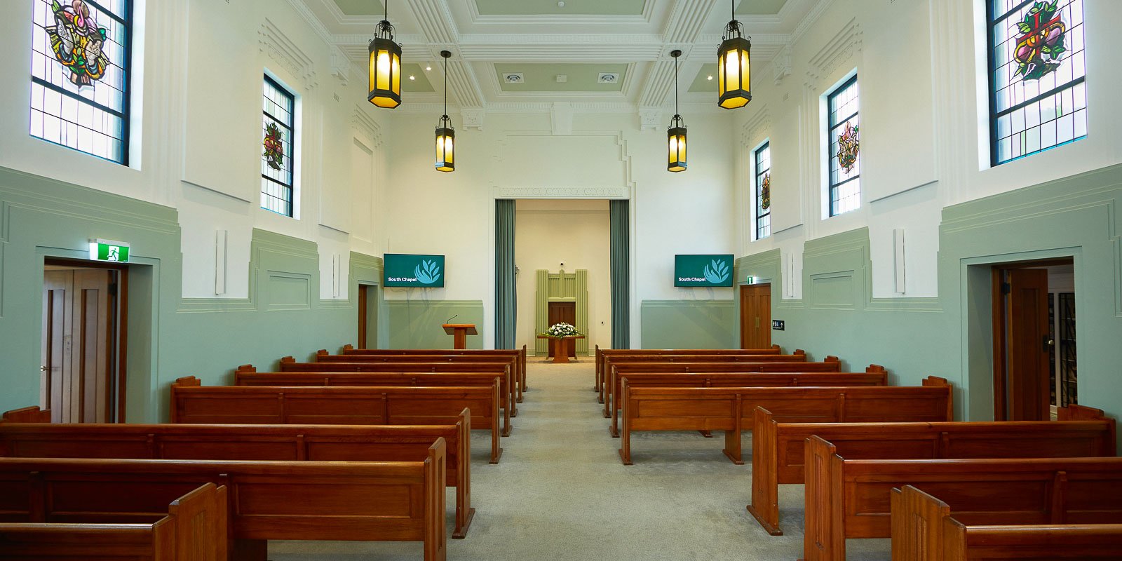 Traditional chapel interior with wooden pews, stained glass windows, and hanging lanterns, featuring a stage at the front.