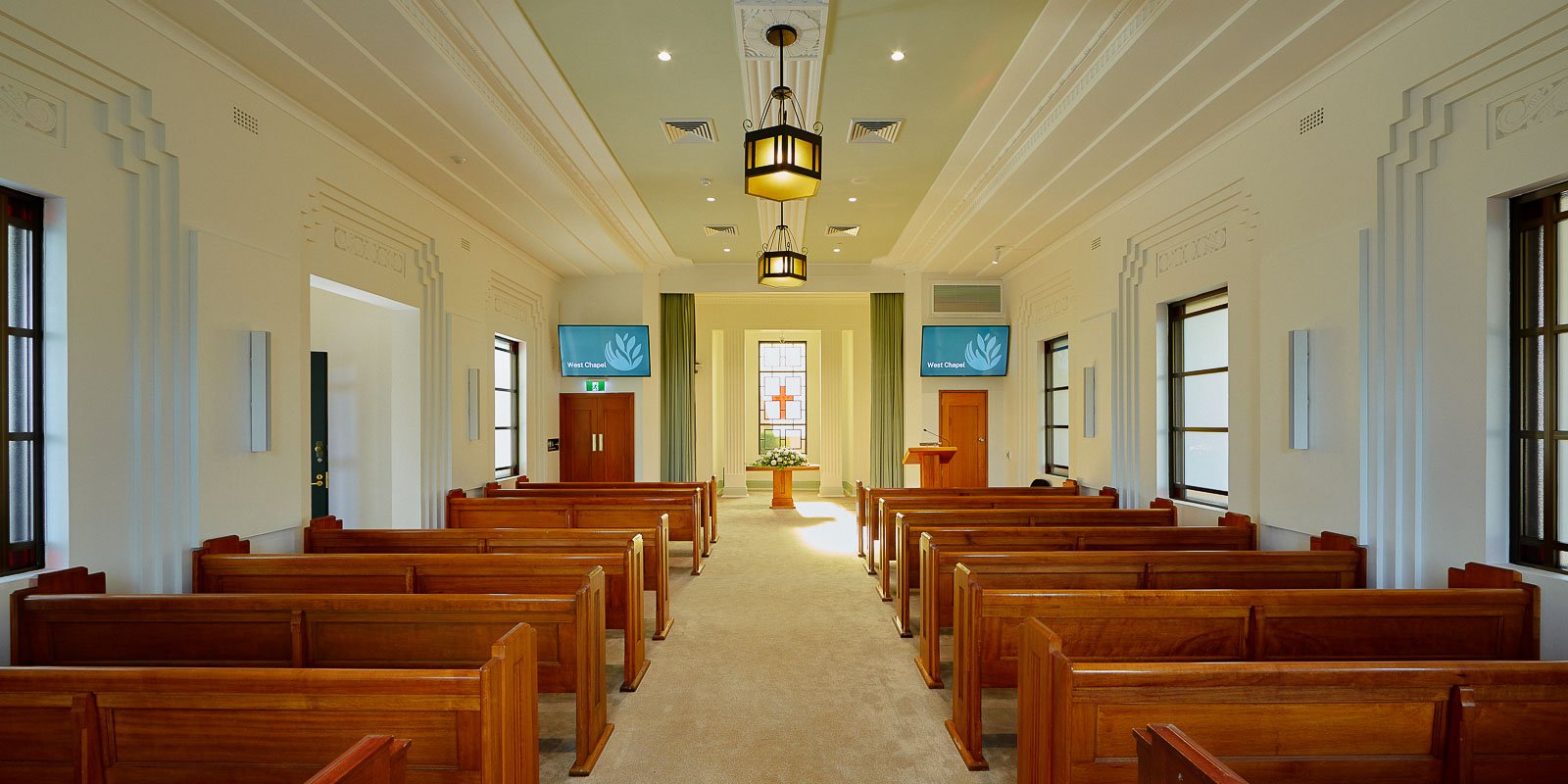 Traditional chapel interior with wooden pews, stained glass windows, and hanging lanterns, featuring a stage at the front.