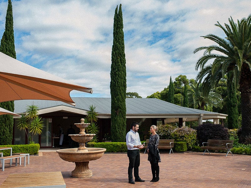 Two people talking near a fountain in a courtyard surrounded by tall trees, with benches and a function building in the background