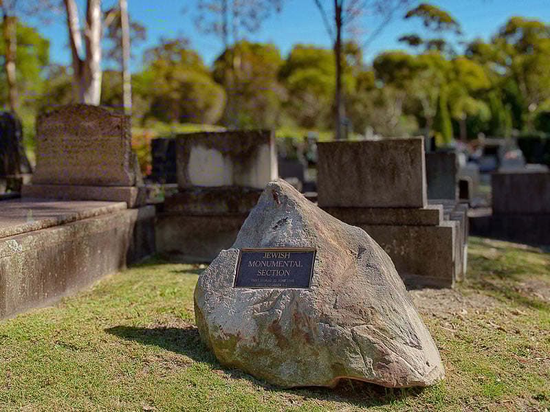 Jewish monumental section in a cemetery, with a memorial plaque on a large stone and several traditional headstones in the background