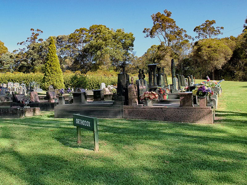 Orthodox monumental section in a cemetery with headstones and flower arrangements, surrounded by trees and greenery.