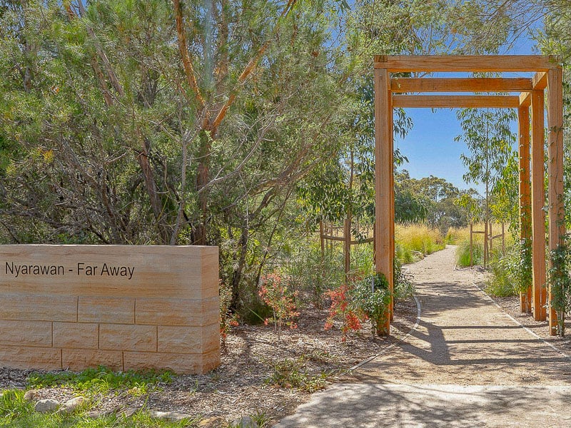 Entrance to a natural burial site with a wooden archway, surrounded by native plants and trees, featuring a sign that reads 'Nyarawan - Far Away