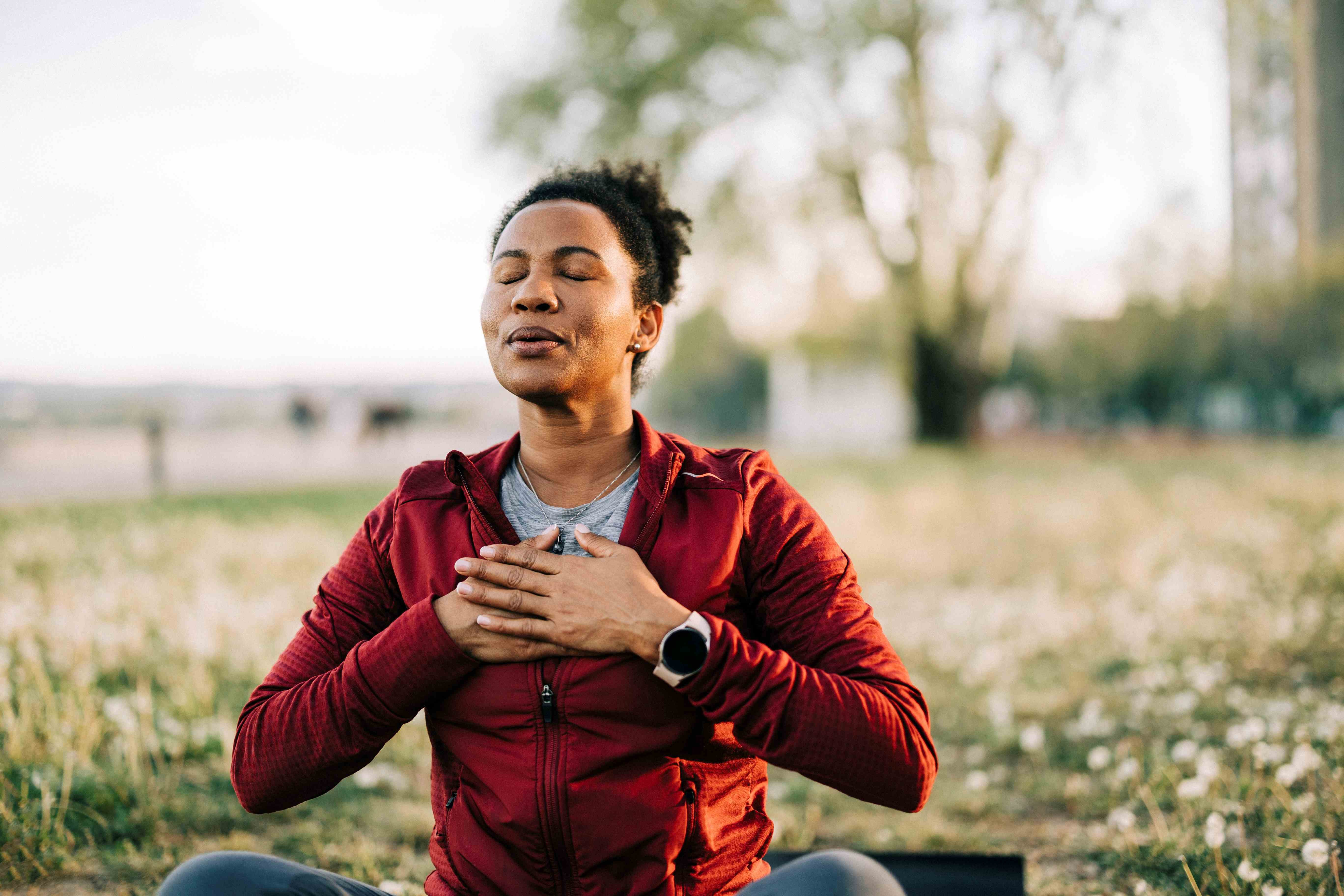 A person meditates outdoors, eyes closed, with hands on their chest, wearing a red jacket. They sit in a field, conveying calm and mindfulness.