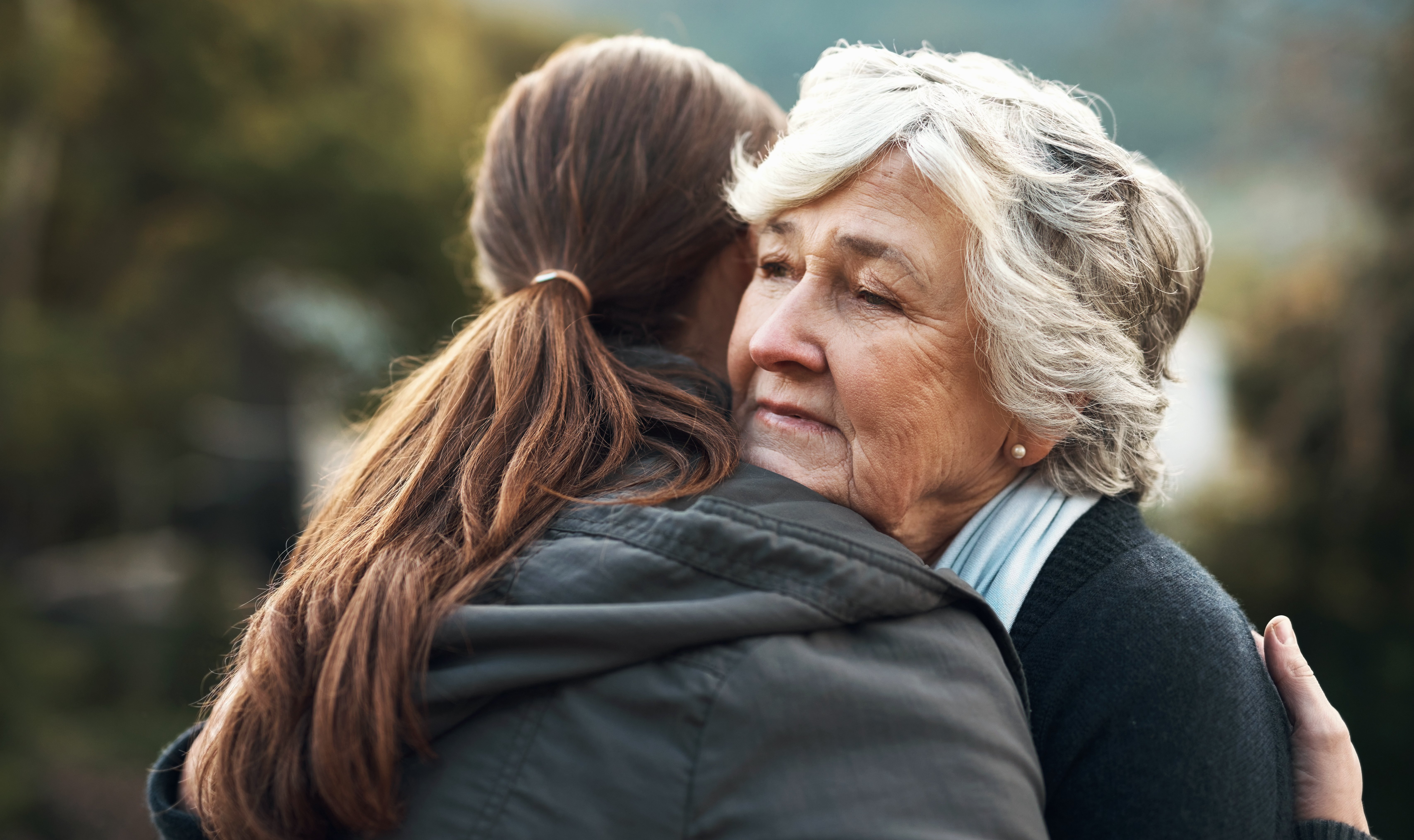 Elderly woman with gray hair hugs a younger person outdoors, conveying comfort and warmth. Her expression suggests a sense of calm and reassurance.