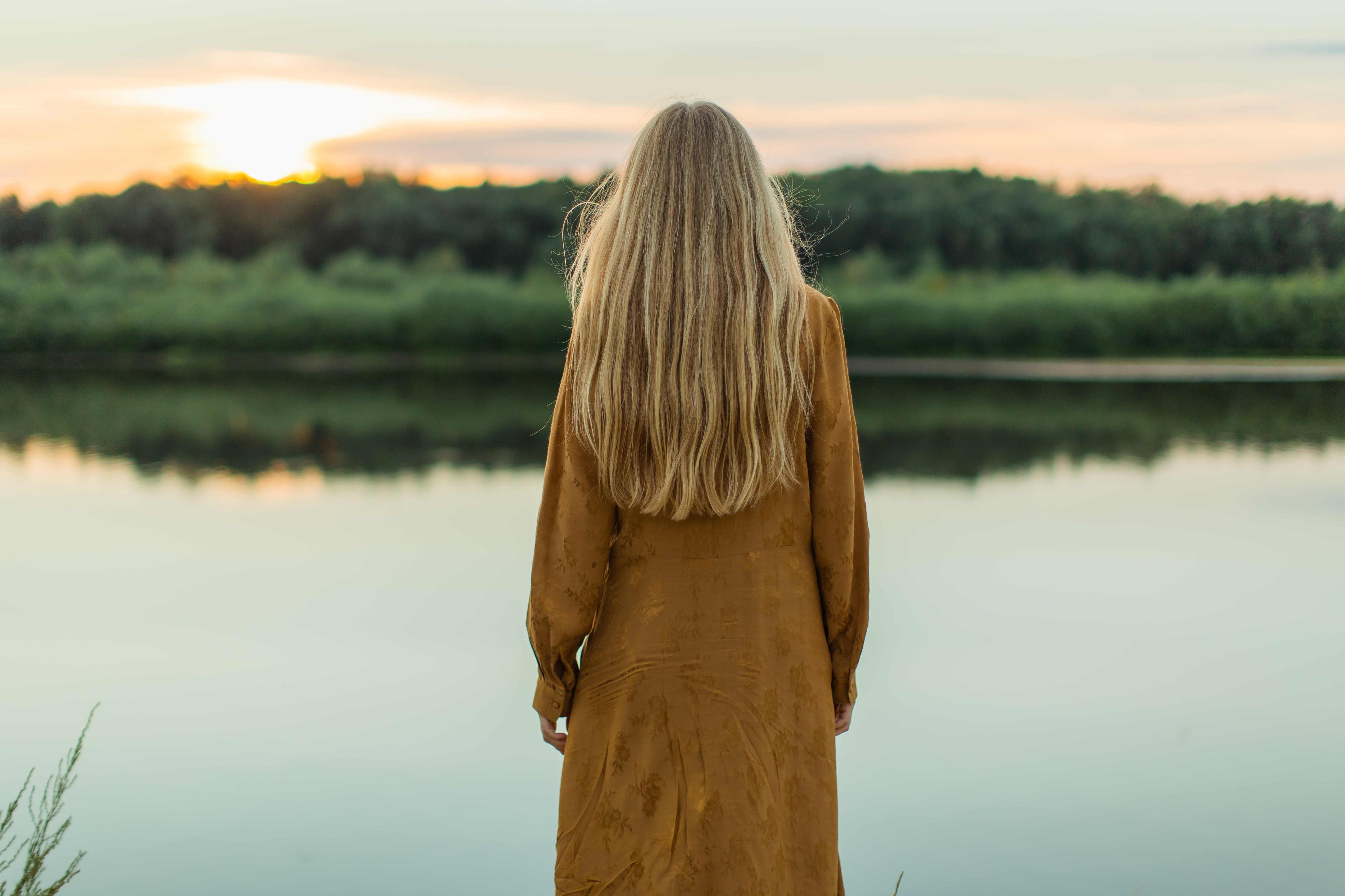 A woman with long blonde hair, wearing a brown dress, stands by a calm lake at sunset. The serene scene conveys a sense of tranquility and introspection.