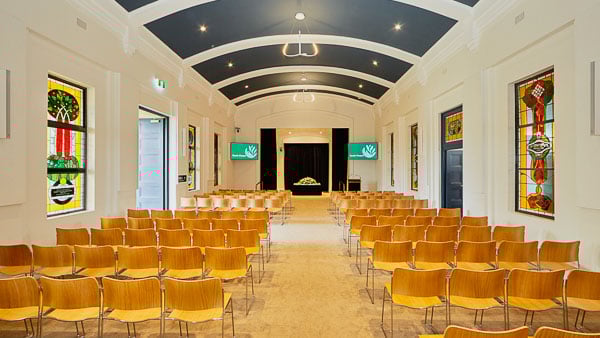 Interior of a chapel with rows of wooden chairs, stained glass windows, and a stage at the front, featuring a ceiling and modern lighting
