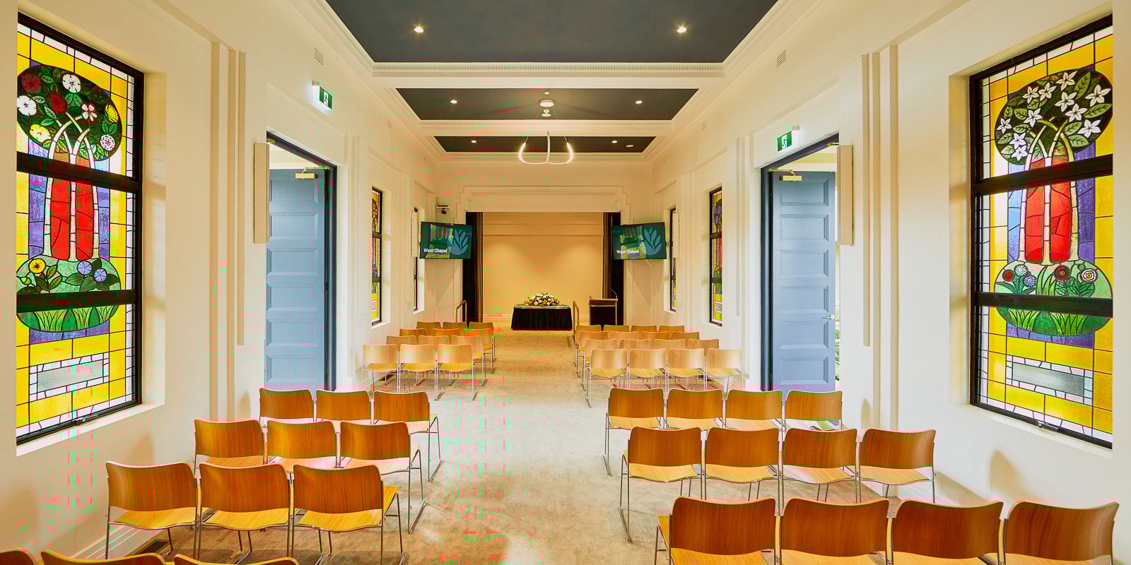 Interior of a chapel with rows of wooden chairs, stained glass windows, and a stage at the front, featuring a ceiling and modern lighting