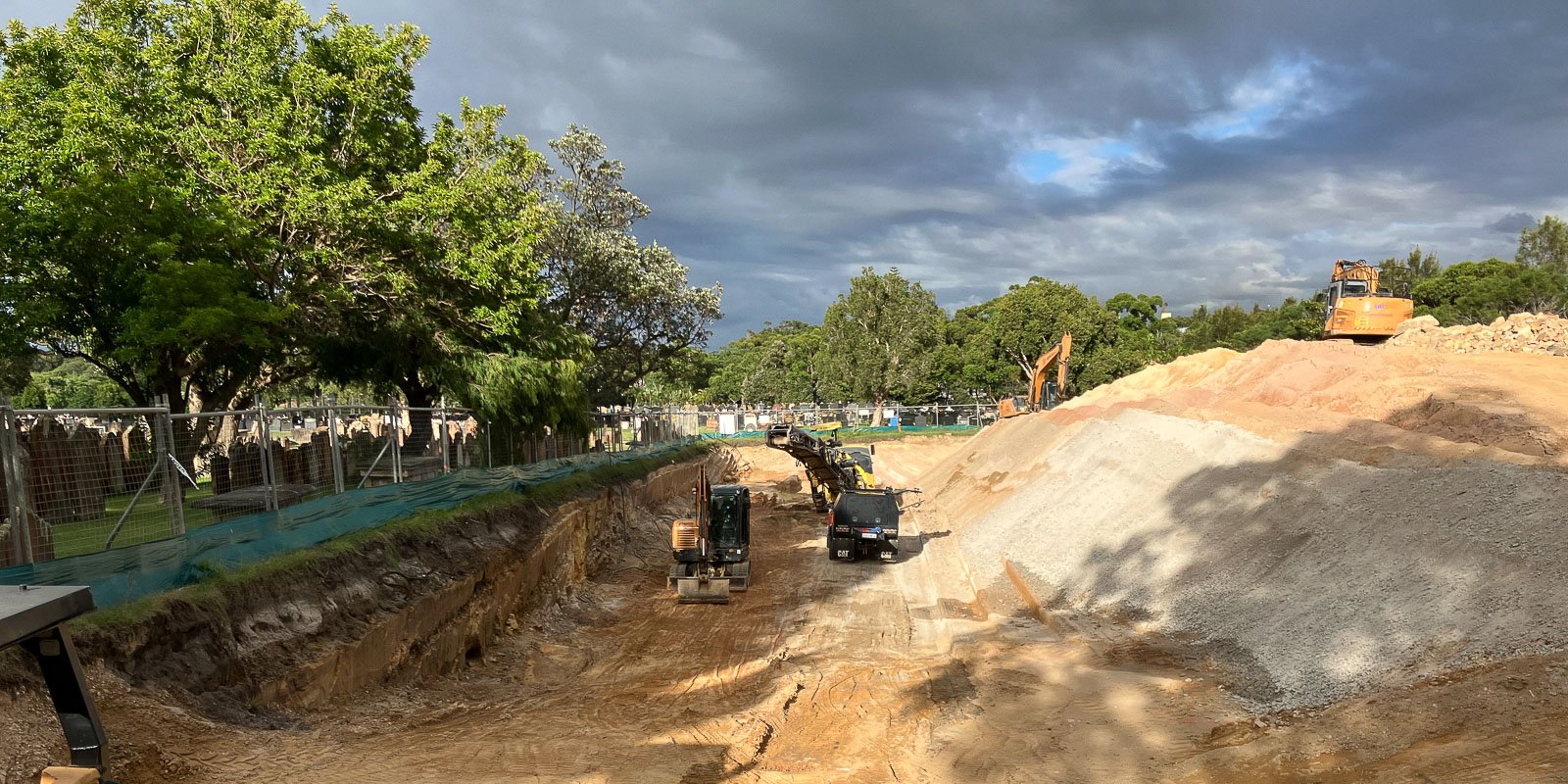 Construction site with excavators and trucks working in a cemetery, with trees and gravestones in the background under a cloudy sky