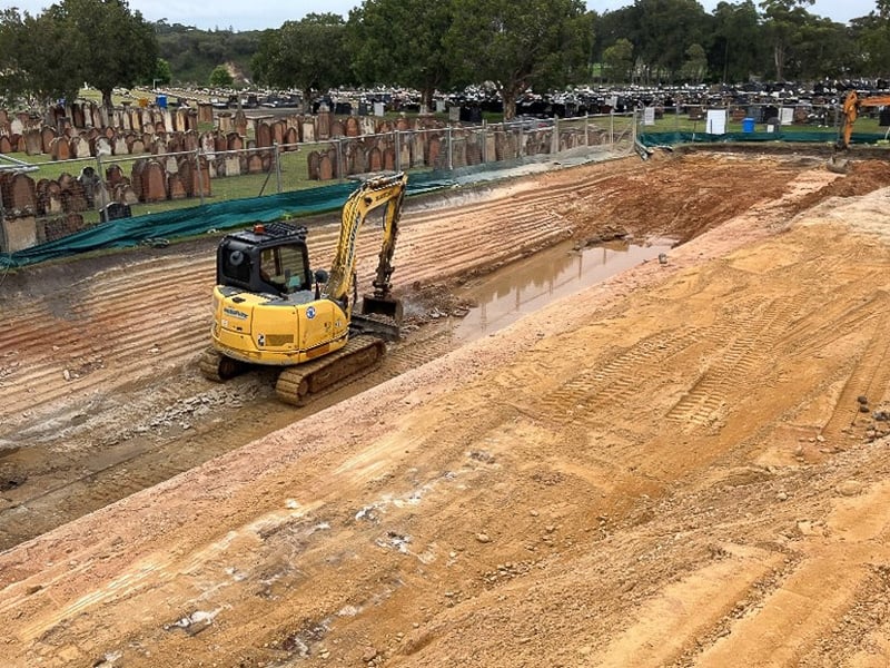 Excavation work in progress at Pioneer Park in Eastern Suburbs Memorial Park with a yellow digger on a cleared site, bordered by heritage gravestones and green fencing in the background