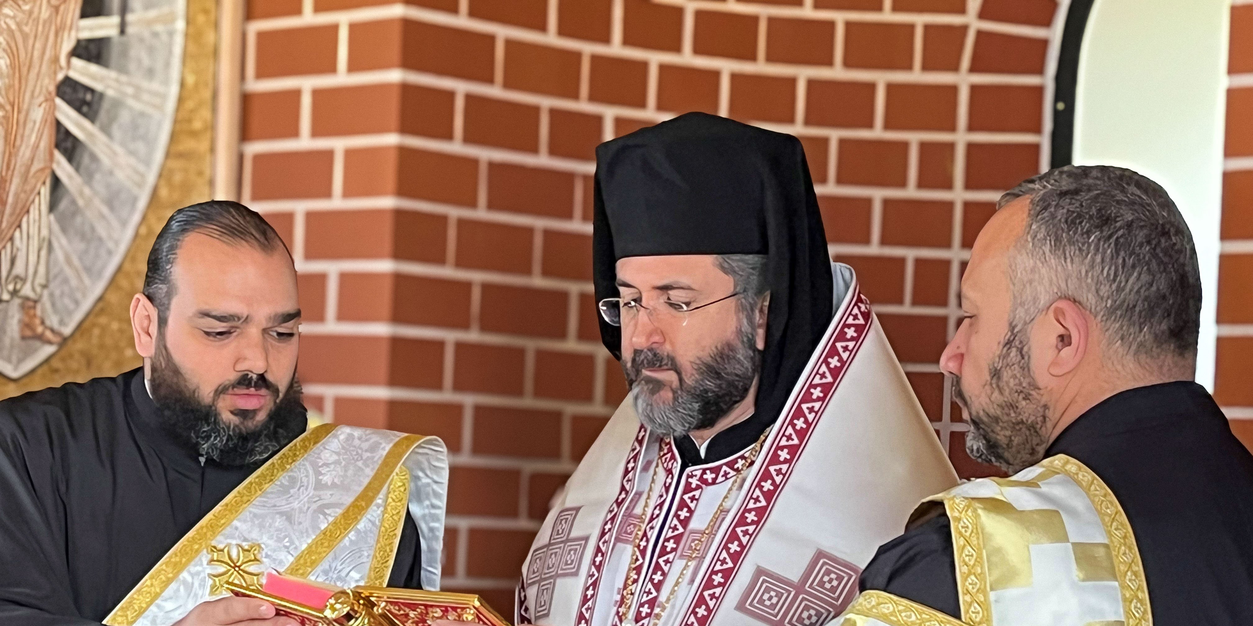 Antiochian Orthodox Archdiocese clergy members in participating in a religious blessing at Rookwood General Cemetery, with a brick wall and religious mosaic in the background