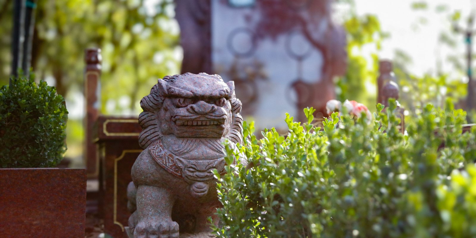 Stone lion statue surrounded by green bushes in a garden at Rookwood Crematorium. The setting is peaceful with sunlight filtering through the trees.