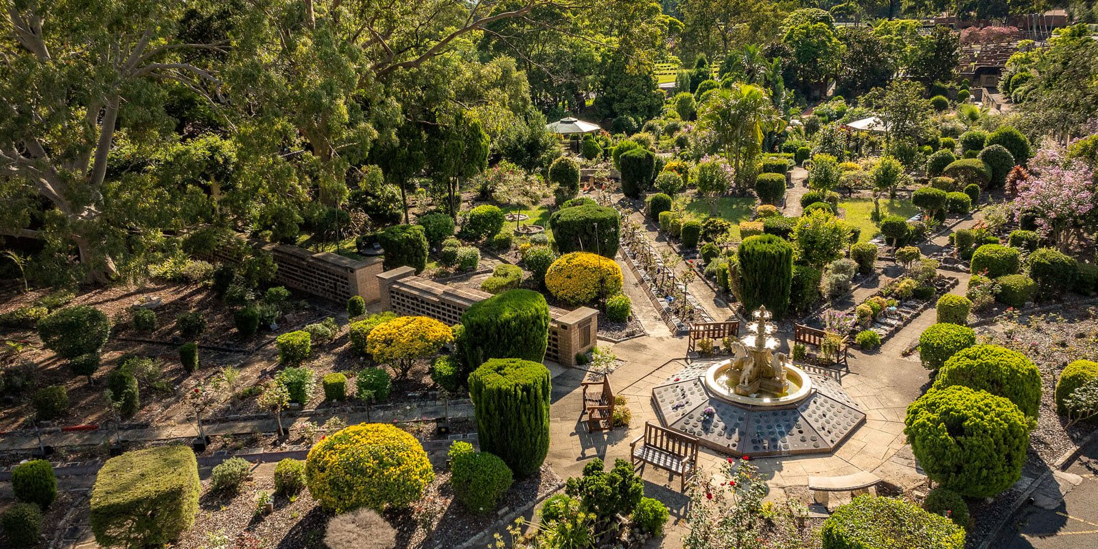 Aerial view of a lush, manicured garden featuring a central fountain, geometric hedges, winding pathways, and shady trees at Rookwood Crematorium.