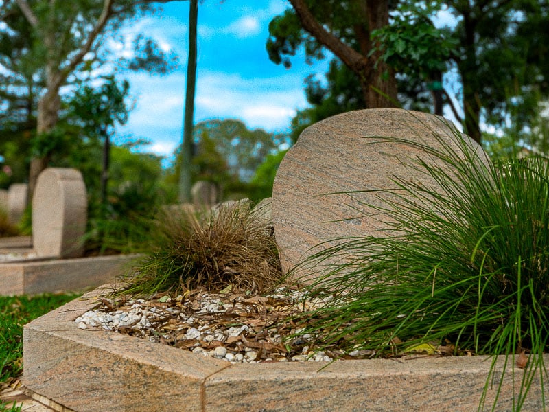Granite octagonal shaped monument surrounded by greenery and stones in a peaceful cemetery. Trees and a blue sky are visible in the background