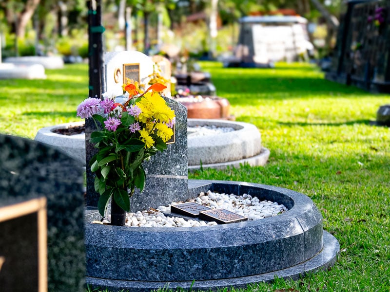 A rounded granite memorial at Rookwood Crematorium surrounded by green grass, adorned with bright flowers and small rocks. Two bronze plaques are placed,